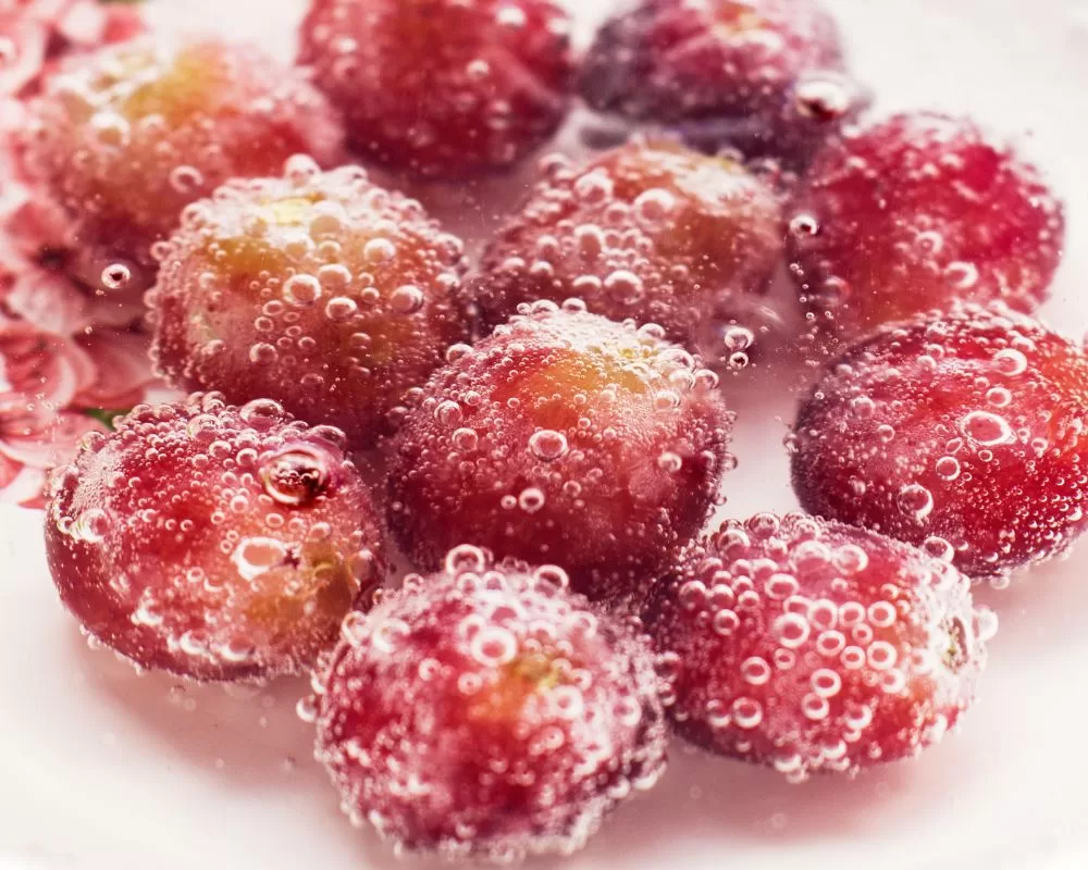 An image showing a cluster of glistening grapes with a pinkish-red hue, partially submerged in water and effervescing with tiny bubbles clinging to their surface, giving a refreshing and tantalizing visual appeal