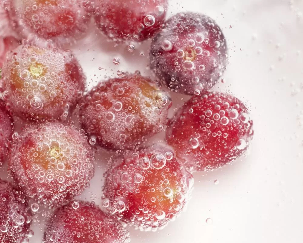 Close-up of fresh red grapes immersed in a carbonated water with multiple small bubbles adhering to their surfaces, highlighting the natural variations in colour from red to purple against a soft white background